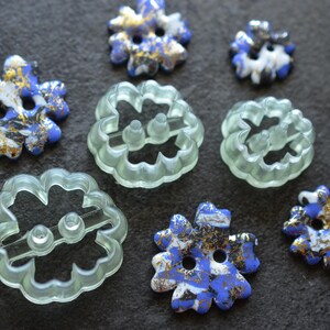 May include: A collection of flower-shaped buttons in two varieties: several opaque buttons featuring a marbled blue, white, and gold pattern, and three translucent, pale green buttons with a scalloped edge and two central attachment holes.