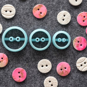 May include: Three blue plastic button cutters of different sizes. The cutters are arranged in a row from largest to smallest. There are many colorful buttons scattered around the cutters.