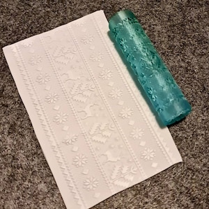 May include: A white silicone baking mat with a raised pattern of snowflakes, trees, and reindeer. A light blue rolling pin with a matching pattern is lying on top of the mat.