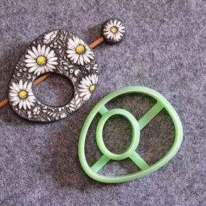 May include: A decorative hair pin with a daisy design and a light green, oval-shaped object. The hair pin features a wooden stick and a black and white daisy design. The green object has an oval center and four connecting bars.