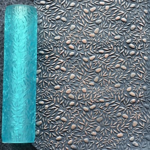 May include: A light blue rolling pin with a raised pattern of leaves and berries. The rolling pin is next to a clear plastic sheet with the same pattern embossed on it.