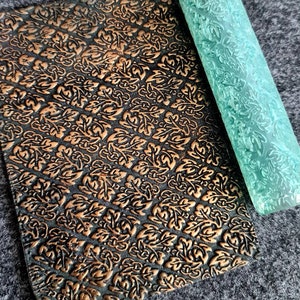 May include: A green rolling pin with a raised floral design. The rolling pin is next to a sheet of embossed paper with a similar floral design.