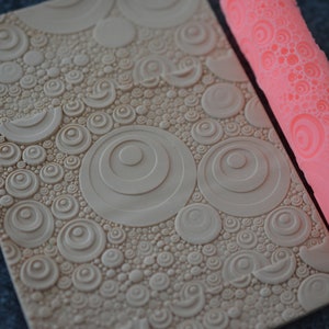May include: A pink silicone rolling pin with a circular pattern, and a beige textured sheet with a similar pattern. The rolling pin is used to create embossed designs on fondant or clay.