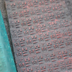 Può includere: Un mattarello di plastica blu con un motivo geometrico. Il mattarello è accanto a un pezzo di materiale grigio scuro con un motivo geometrico rosso e grigio.