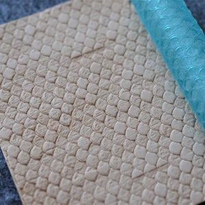 May include: A light blue rolling pin with a textured surface, designed to create a scale pattern on fondant or clay. The rolling pin is lying on a beige surface with a scale pattern.