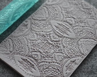 May include: A gray silicone rolling pin with an intricate, detailed pattern. The pattern is a repeating design of swirls and curves.
