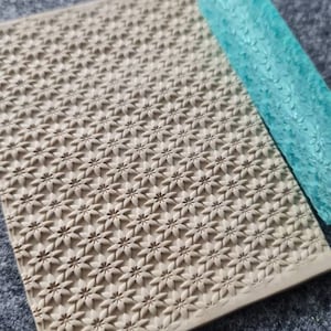 May include: A beige silicone rolling pin with a repeating star pattern. The rolling pin is on a gray surface.