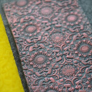 Può includere: Un rullo di gomma giallo con un motivo dettagliato, accanto a un foglio color rame con un motivo in rilievo ripetuto di intricati disegni floreali.
