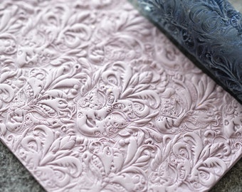 May include: A close-up of a light pink textured surface with a detailed floral pattern. A dark blue rolling pin with a similar floral pattern is lying on top of the surface.