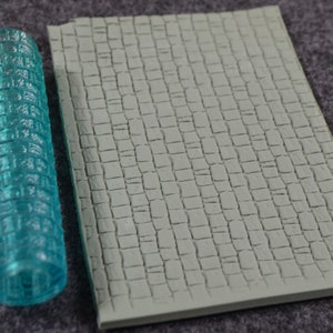 May include: A clear, cylindrical roller with a textured brick pattern, next to a rectangular, light green mat with a cobblestone design. The roller is a translucent teal color.