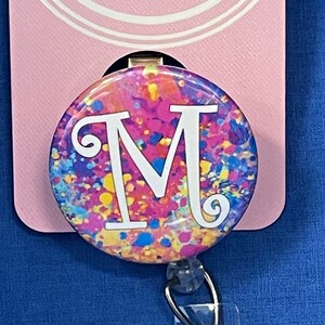 May include: A colorful badge reel with a white letter "M" on a pink, yellow, blue, and green splatter background. The reel is attached to a clear plastic badge holder with a silver clip. The card behind the reel says "That's My Style".