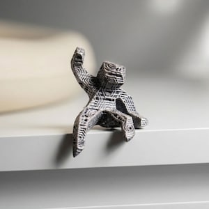 May include: A small, grey figurine of a stylized figure with an arm raised. The figurine has a textured surface with geometric patterns. It is sitting on a white surface.