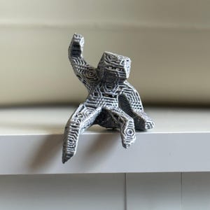 May include: A grey, 3D-printed sculpture of a figure with an outstretched arm. The figure is sitting on a white surface. The sculpture has a geometric, web-like design throughout its form.