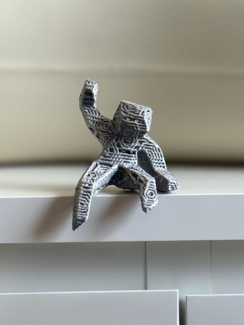May include: A grey, 3D-printed sculpture of a figure with an outstretched arm. The figure is sitting on a white surface. The sculpture has a geometric, web-like design throughout its form.