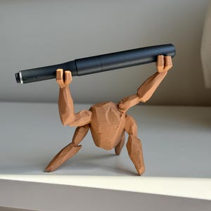May include: A brown, 3D-printed pen holder shaped like a person holding a black pen. The pen holder has articulated limbs and a textured surface. The pen is dark grey with silver detailing. The pen holder is approximately 15 cm long.