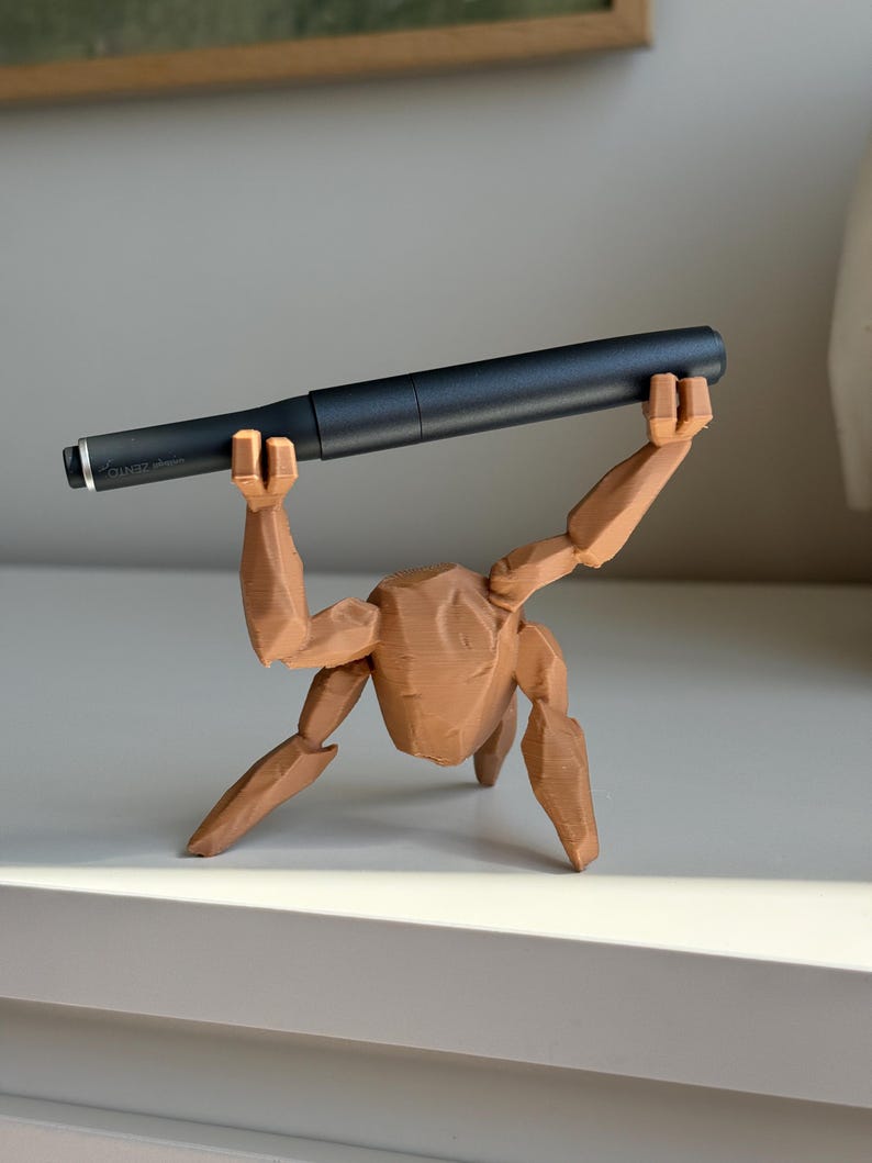 May include: A brown, 3D-printed pen holder shaped like a person holding a black pen. The pen holder has articulated limbs and a textured surface. The pen is dark grey with silver detailing. The pen holder is approximately 15 cm long.