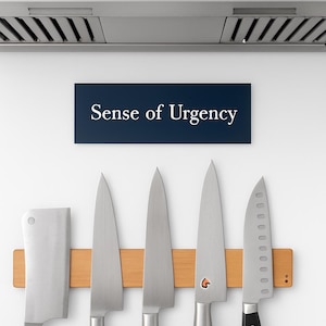 May include: A set of five stainless steel kitchen knives on a wooden magnetic knife holder. The knives are displayed against a white wall, with a stainless steel range hood above. A sign reads "Sense of Urgency".