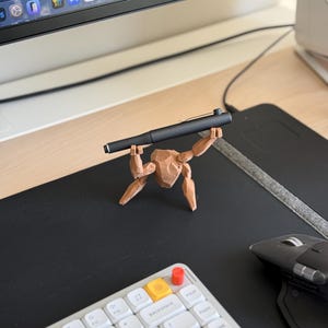 May include: A black pen held by a small, brown, wooden figurine. The figurine is posed as if lifting the pen. The pen and figurine are on a black desk mat, with a white keyboard and computer monitor in the background.