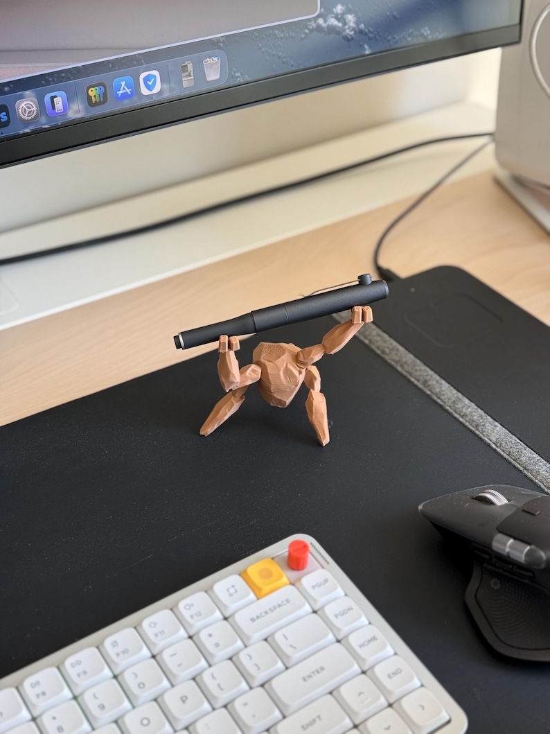 May include: A black pen held by a small, brown, wooden figurine. The figurine is posed as if lifting the pen. The pen and figurine are on a black desk mat, with a white keyboard and computer monitor in the background.