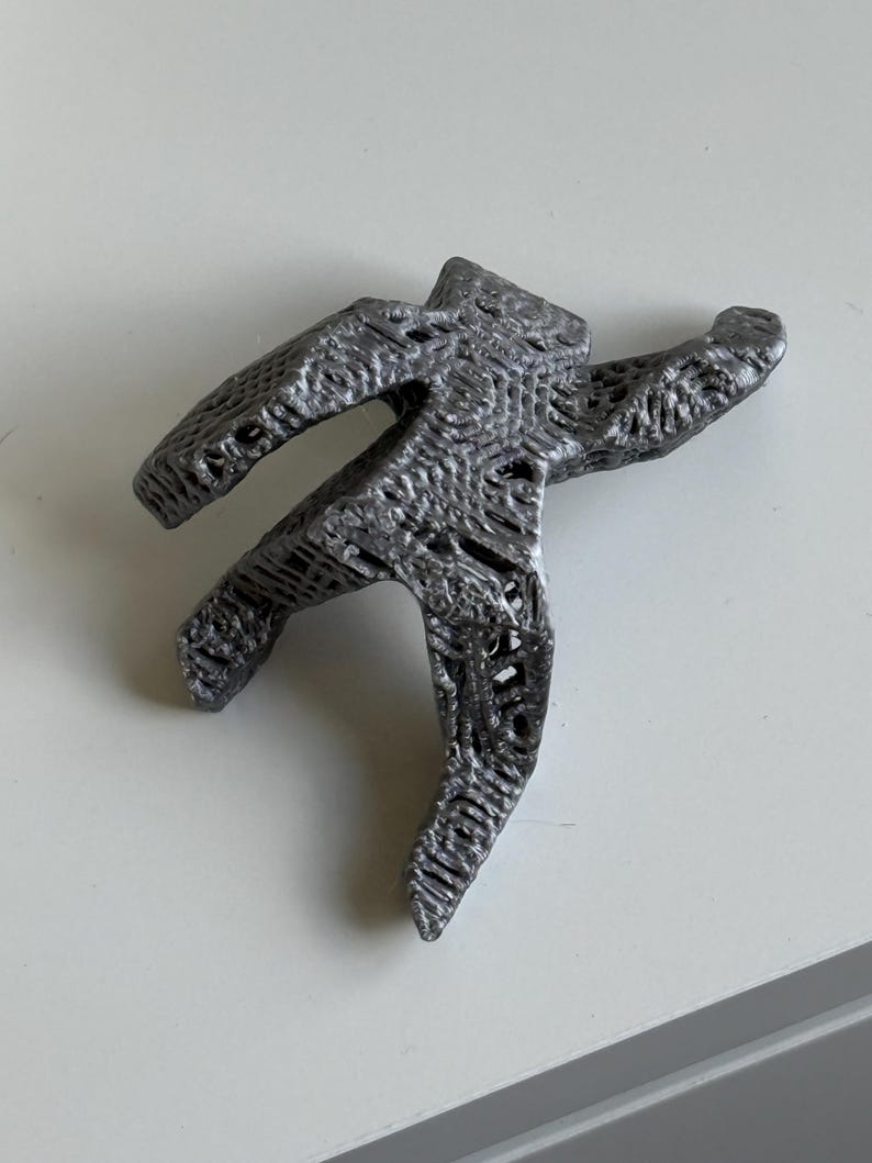 May include: A grey, 3D-printed sculpture of a person in a star shape, with arms and legs outstretched. The sculpture has a textured surface and is placed on a white surface.