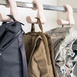 May include: Three backpacks hanging on a metal rod with pink hooks. The backpacks are in shades of black, brown, and gray. The hooks are designed to hold the backpack straps securely.