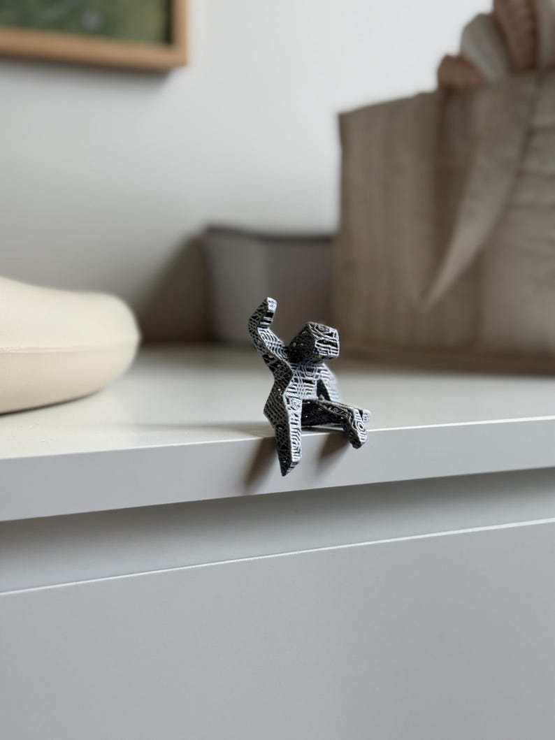 May include: A small, abstract sculpture of a person sitting on a ledge. The figure is black and white with a geometric pattern. The sculpture is perched on the edge of a white surface.