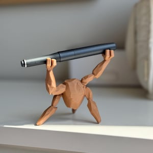 May include: A dark grey pen is held aloft by a brown wooden figurine. The pen has a silver tip and a modern design. The figurine is in a lifting pose, adding a unique touch to the pen display. The pen is a writing instrument.