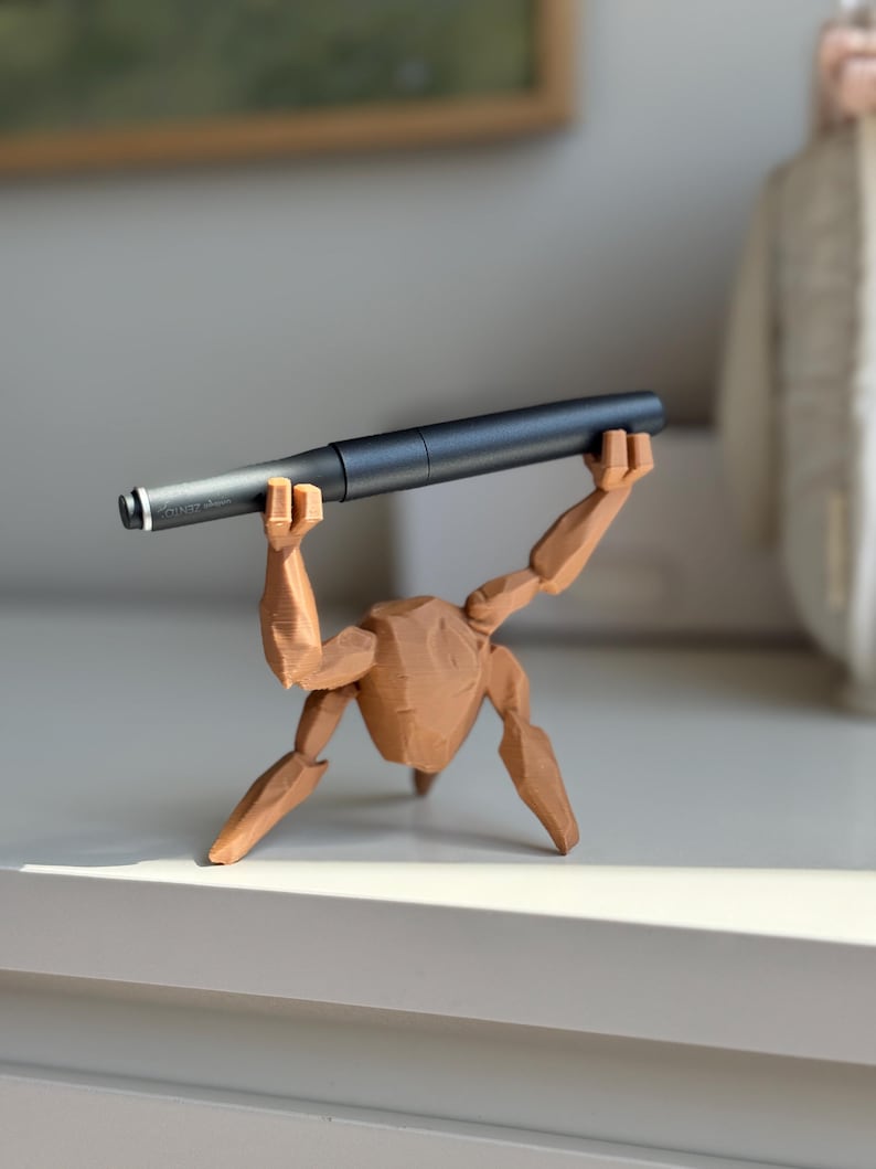 May include: A dark grey pen is held aloft by a brown wooden figurine. The pen has a silver tip and a modern design. The figurine is in a lifting pose, adding a unique touch to the pen display. The pen is a writing instrument.