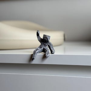 May include: A small, detailed figurine of a person in a seated position, with one arm raised. The figurine is black and white, with intricate patterns covering its surface. It is perched on the edge of a white surface.