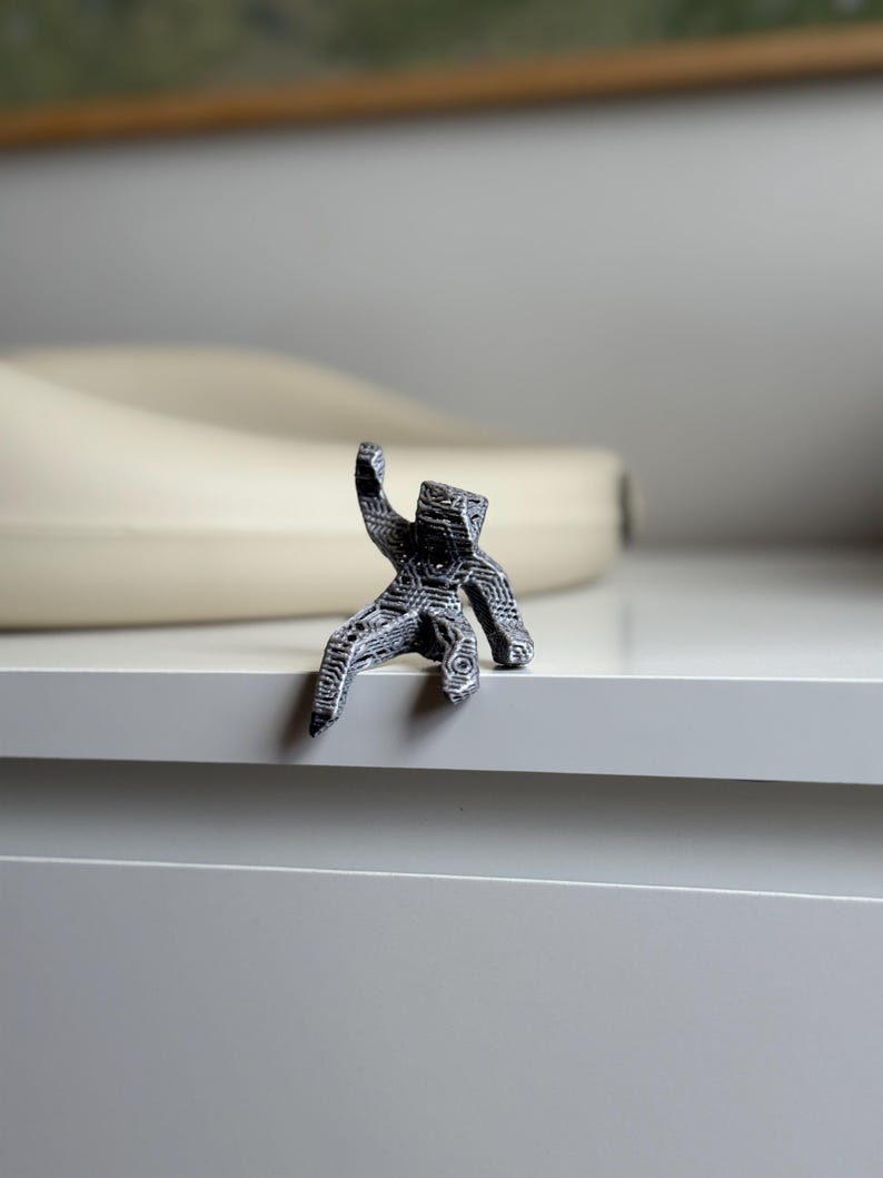 May include: A small, detailed figurine of a person in a seated position, with one arm raised. The figurine is black and white, with intricate patterns covering its surface. It is perched on the edge of a white surface.