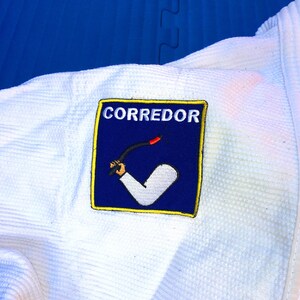 May include: White martial arts uniform with a blue and yellow patch that says "CORREDOR" and depicts a black stick being held by a hand with a white sleeve.
