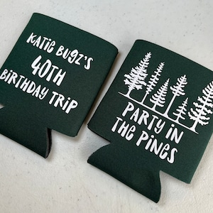 May include: Two green foam can coolers with white text. One cooler says "Katie Bugz's 40th Birthday Trip". The other cooler says "Party in the Pines" and has a design of five white pine trees.