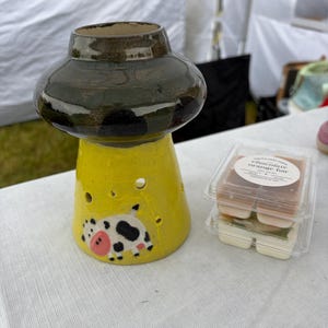 May include: A ceramic wax warmer with a dark green mushroom-shaped top and a bright yellow base featuring a painted black and white cow with a pink nose. Next to it are two stacked clear plastic containers of wax melts labeled chocolate orange bar.