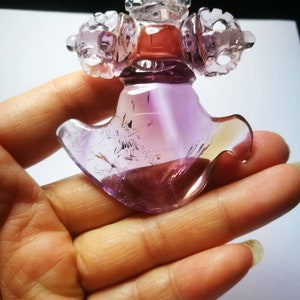 May include: A purple amethyst crystal carved into a detailed, intricate design. The crystal is translucent and has a light purple hue. The carving is of a traditional Tibetan Buddhist symbol.