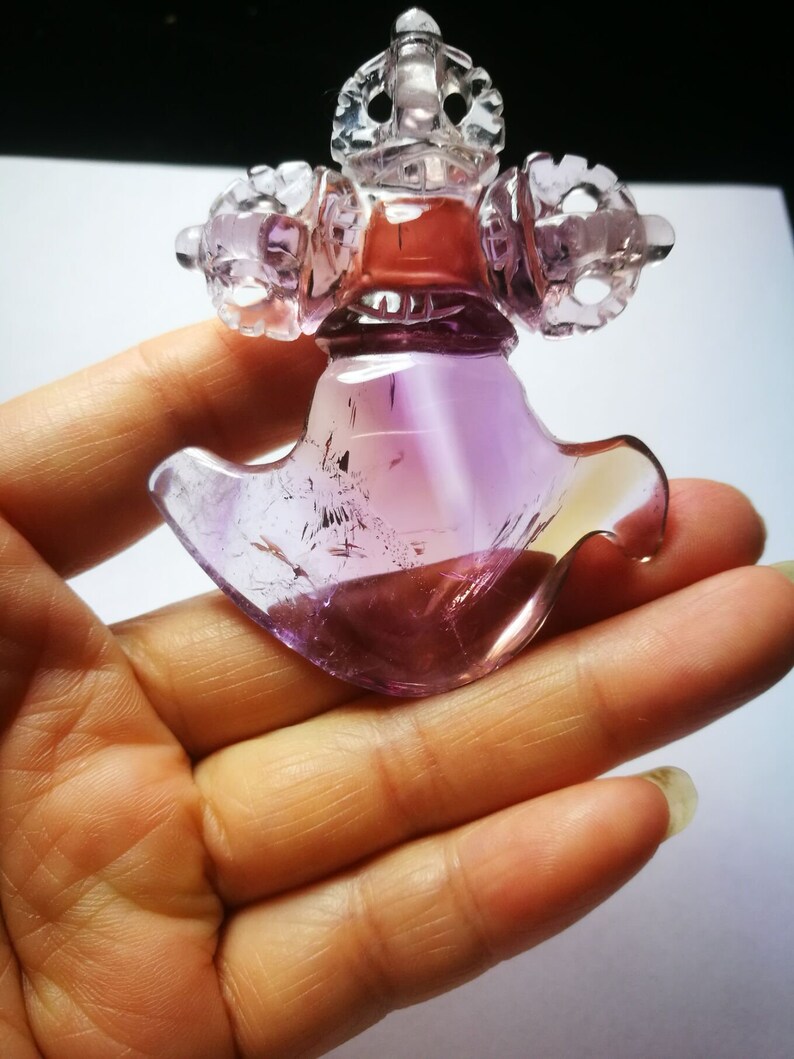 May include: A purple amethyst crystal carved into a detailed, intricate design. The crystal is translucent and has a light purple hue. The carving is of a traditional Tibetan Buddhist symbol.