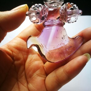 May include: A purple and clear glass sculpture of a stylized figure with intricate details. The figure has a crown-like top and a flowing, wing-like bottom.