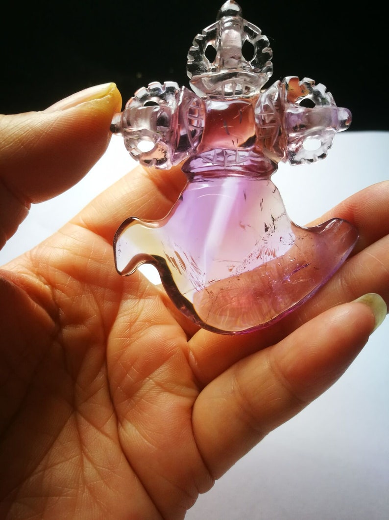 May include: A purple and clear glass sculpture of a stylized figure with intricate details. The figure has a crown-like top and a flowing, wing-like bottom.