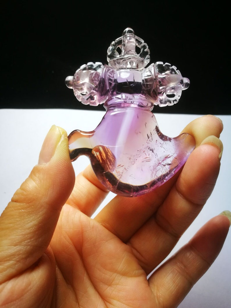 May include: A purple and clear crystal carved into the shape of a Tibetan prayer wheel. The prayer wheel is intricately detailed and has a crown on top.
