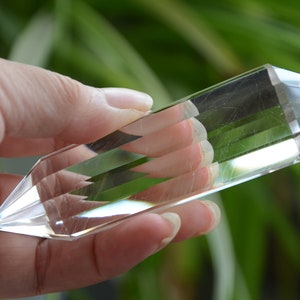 Op de afbeelding: Een heldere kwartskristalpunt met facetgeslepen zijkanten en puntige uiteinden. De kristal wordt in een hand gehouden tegen een vage groene achtergrond. Dit kristal is een decoratief item.