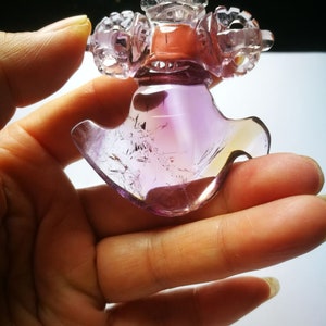 May include: A purple and clear amethyst crystal carved into a detailed, intricate design. The crystal is shaped like a bird with a crown on its head. The crystal is held in a hand.