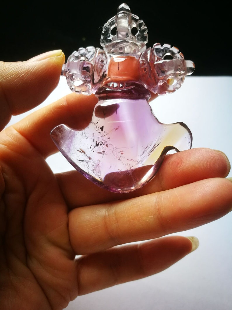 May include: A purple and clear amethyst crystal carved into a detailed, intricate design. The crystal is shaped like a bird with a crown on its head. The crystal is held in a hand.
