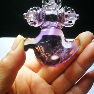 May include: A purple amethyst crystal carved into a detailed, intricate design. The crystal is shaped like a stylized bird with a crown on top. The crystal is translucent and has a light purple hue.