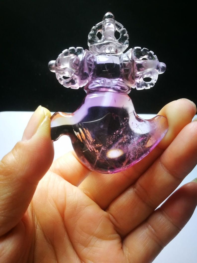 May include: A purple amethyst crystal carved into a detailed, intricate design. The crystal is shaped like a stylized bird with a crown on top. The crystal is translucent and has a light purple hue.