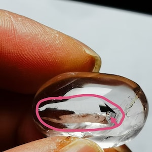 Op de afbeelding: Een helder kwartskristal met een natuurlijke insluiting, een kleine, donkere vlek, zichtbaar in het kristal. Het kristal is glad en gepolijst, met een afgeronde vorm.
