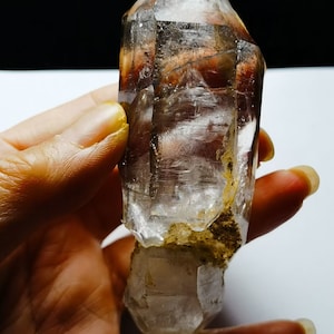 May include: A large, clear quartz crystal with a double termination. The crystal has a natural, rough base and is partially covered in a brown, earthy material.