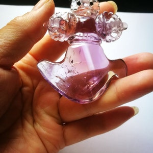 May include: A purple amethyst crystal carved into a detailed, intricate design. The crystal is shaped like a cross with a pointed bottom and has a smooth, polished finish.