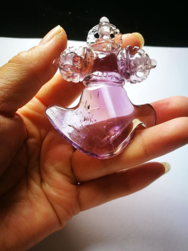 May include: A purple amethyst crystal carved into a detailed, intricate design. The crystal is shaped like a cross with a pointed bottom and has a smooth, polished finish.