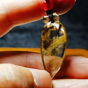 May include: A clear, teardrop-shaped pendant with golden, hair-like inclusions. The pendant is suspended from a black cord with a red bead. The pendant is held between fingers, showcasing its polished surface and intricate internal patterns.