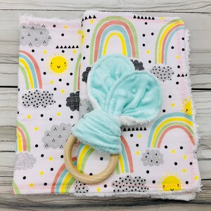 May include: A set of two rainbow-themed baby burp cloths with a light blue teething ring with a wooden ring. The burp cloths have a white background with black polka dots, gray clouds, yellow stars, and multi-colored rainbows.