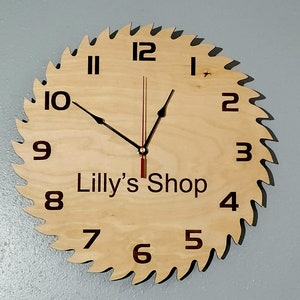 May include: A wooden clock shaped like a saw blade with black hands and Roman numeral markings. The clock face is light brown and has the text "Lilly's Shop" printed on it.